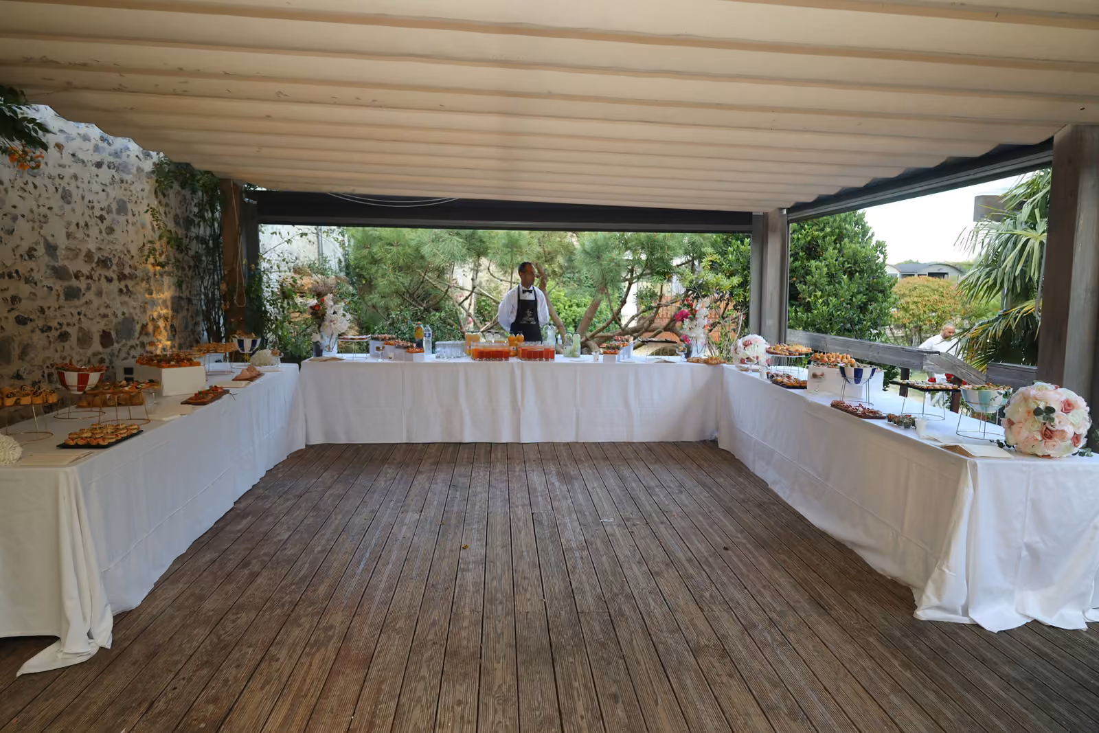 Buffet en U sous une pergola, nappes blanches et parquet en bois, jardin en arrière-plan