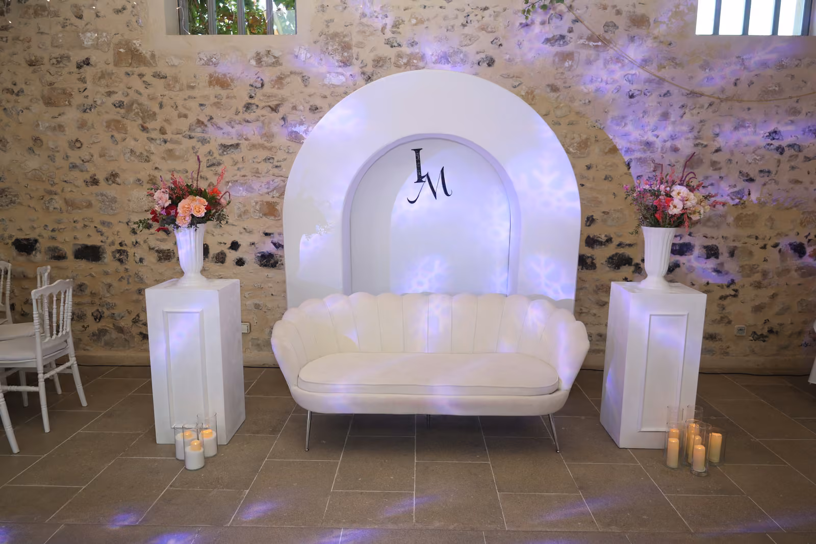 Coin photo des mariés : banquette blanche devant une arche lumineuse aux initiales, fleurs et bougies