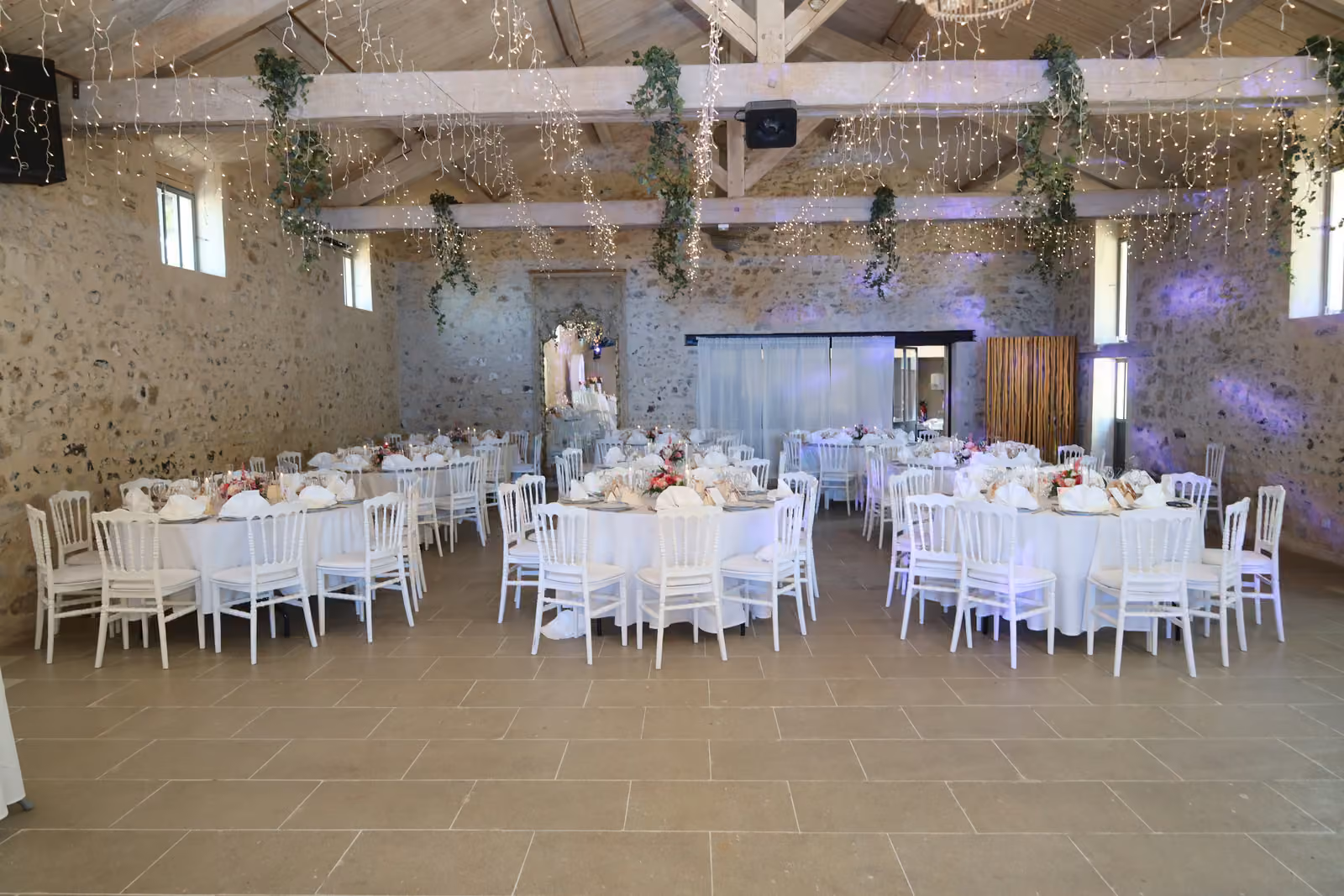 Salle de mariage avec tables rondes nappées de blanc et chaises Napoléon blanches sous une guirlande lumineuse